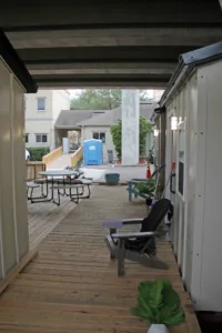 View of tiny home village community from under overpass, with Adirondack chair in foreground and a similar chair and picnic table in background. Across from village in background is a beige-colored building with a blue portable restroom and a wooden ramp leading up to entrance