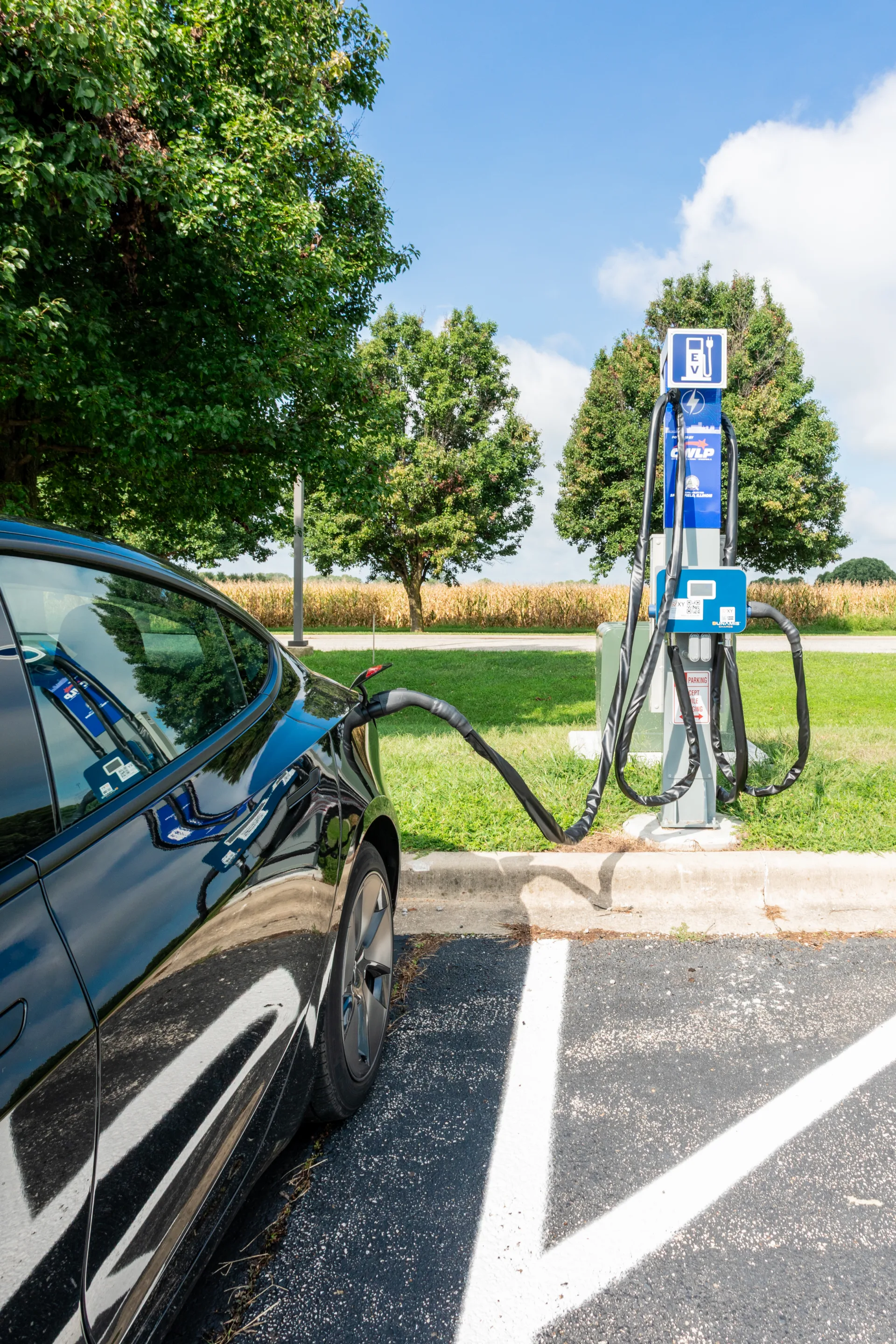 EV charger in background at right; black car at left with charger plugged in; cornfields and trees in background