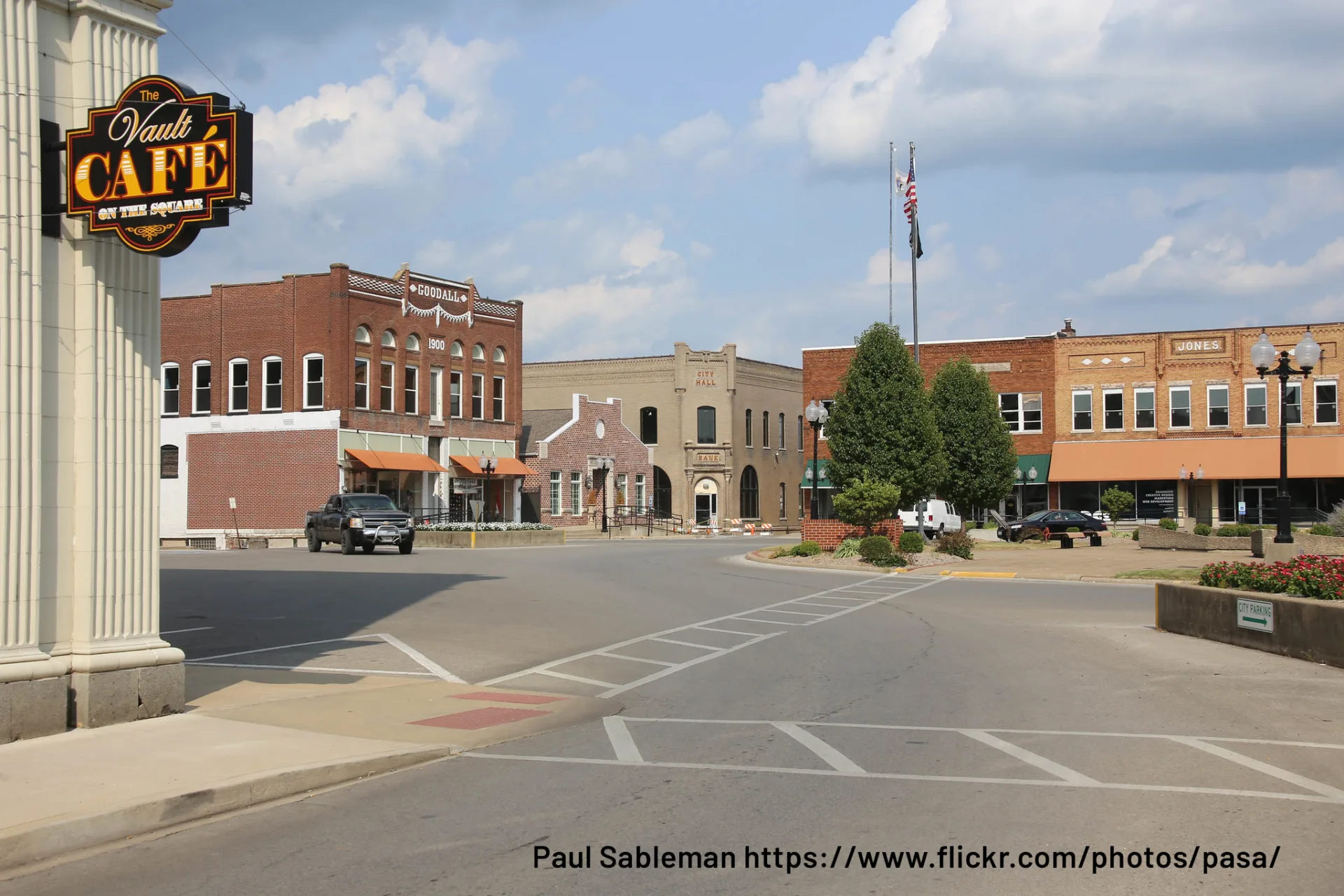 Small-town downtown area with connected buildings and a few cars; building at far left with sign: The Vault Cafe on the Square