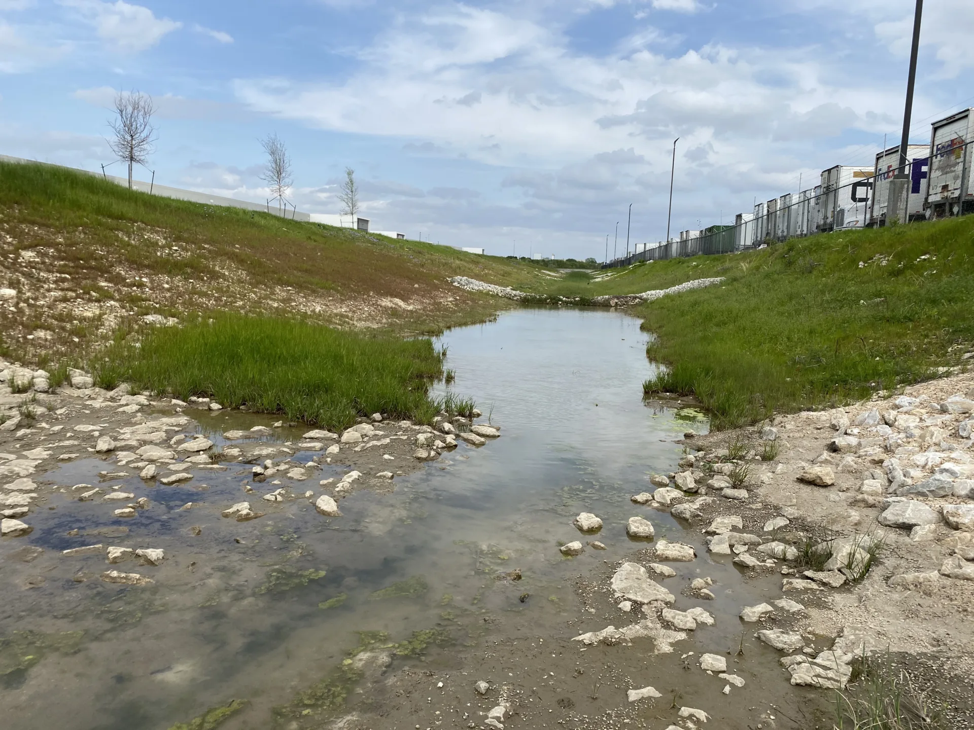 Retention pond in foreground with blue sky and clouds in background and trucks at right