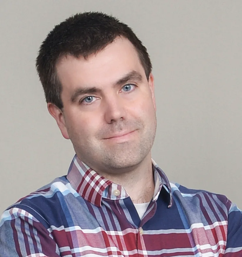 Headshot of Tristan Rickett wearing blue and red plaid button down shirt with white tee underneath, tilting head to left, smiling. Solid beige background