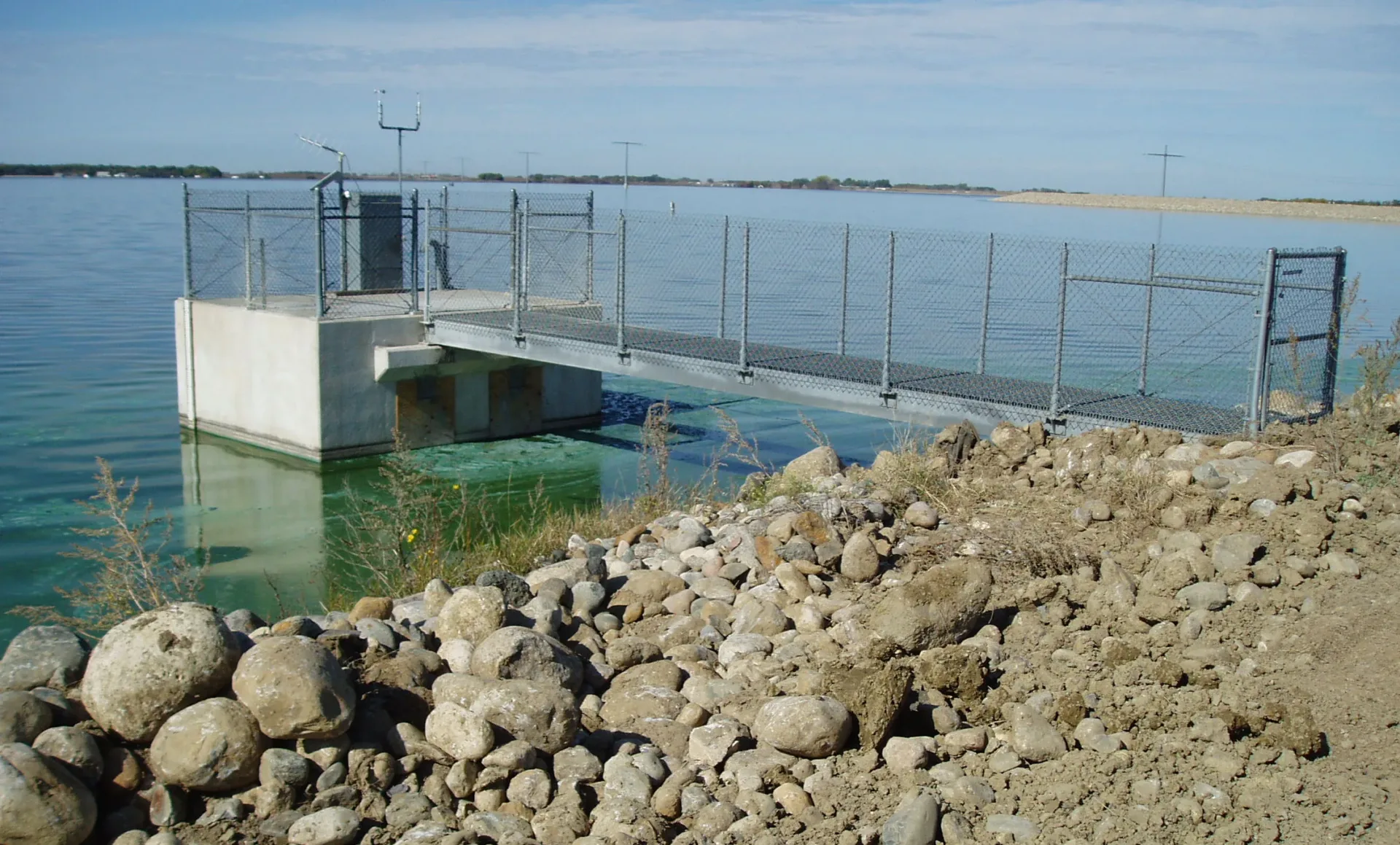 Ground photo of walkway over water leading to a small platform surrounded by a fence; rocks and gravel in the foreground and lake in the background