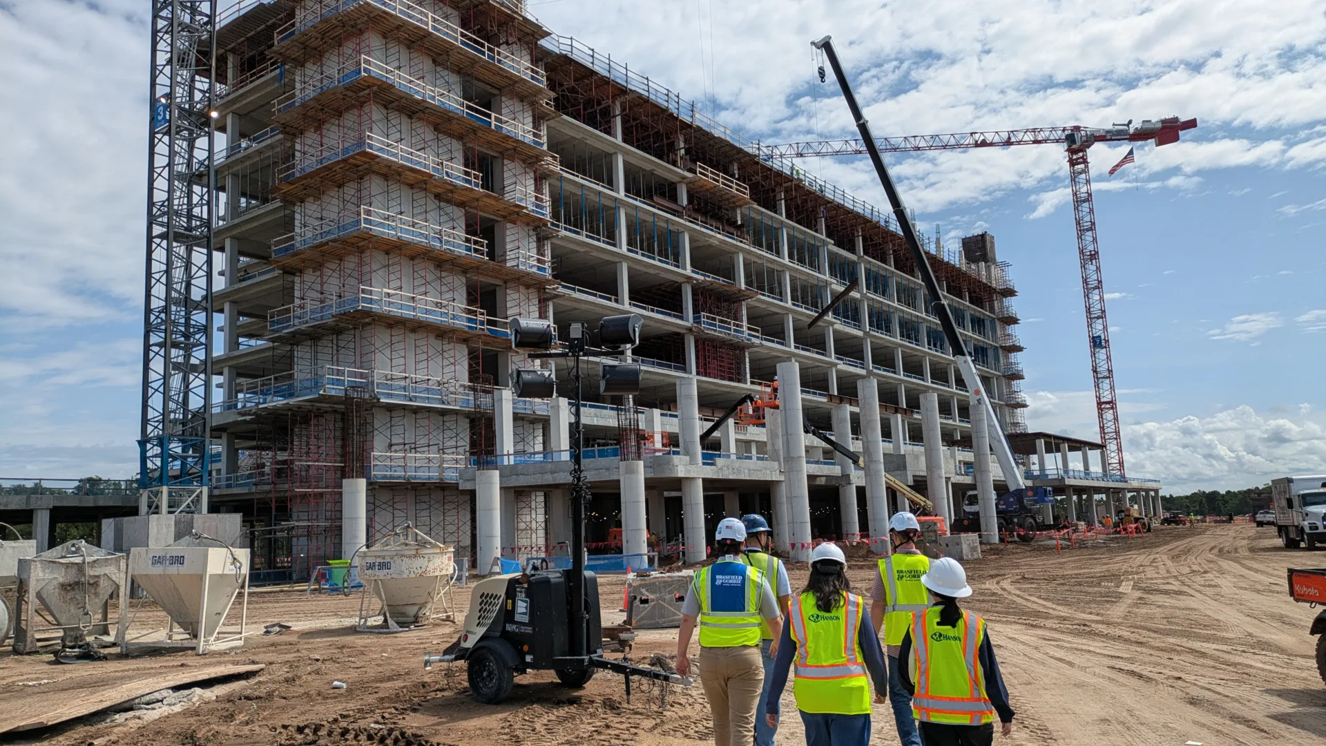 Exterior photo of a hospital building under construction, surrounded by dirt and with cranes on either side; blue sky with puffy white clouds in background; workers in neon-yellow vests and hard hats in foreground