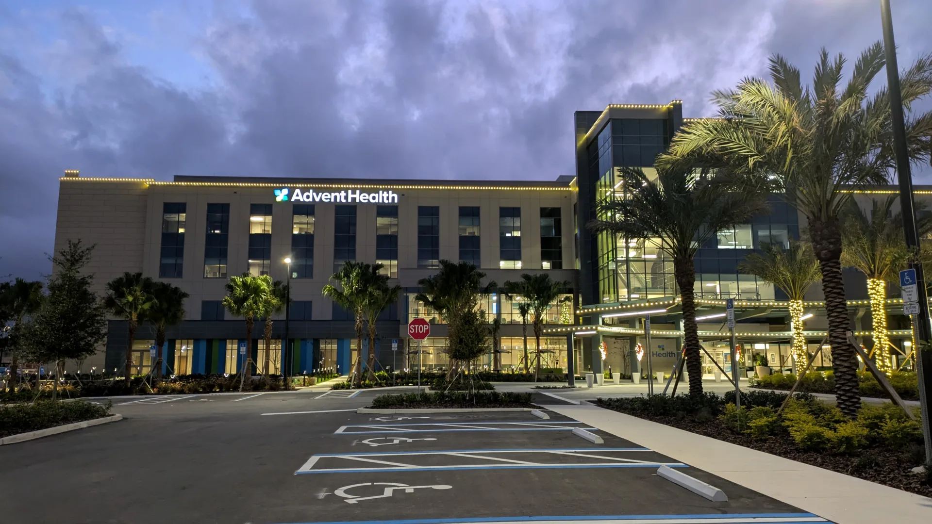 Nighttime exterior photo of front of hospital building with lit-up AdventHealth sign toward top of building, lights around top and around entrance canopy, and palm trees at right