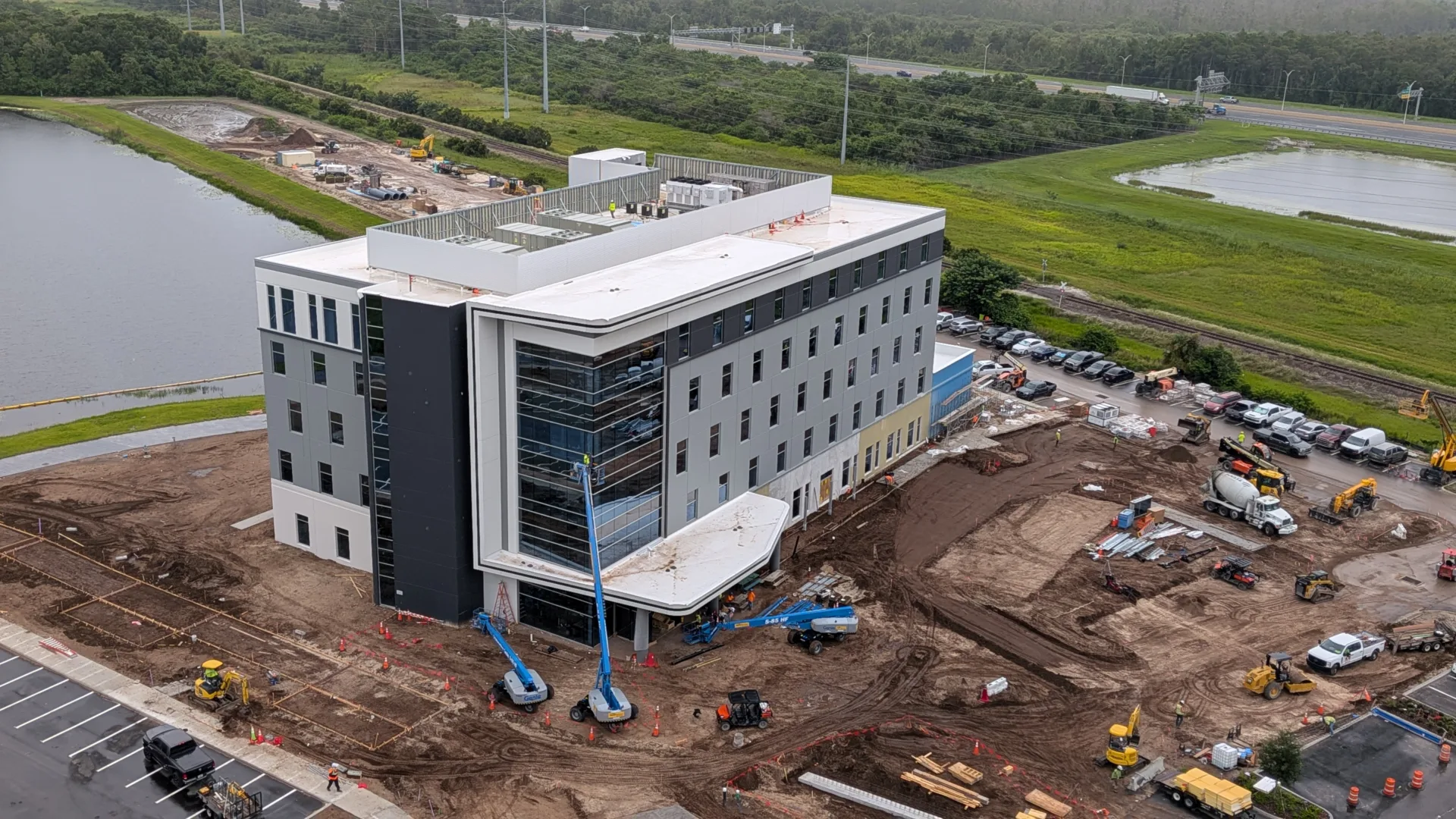 Aerial photo of a medical office building under construction near a pond, surrounded by dirt, cars and construction equipment, with green grass and trees and a roadway in the distance