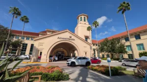Exterior view of Spanish-style hospital building with red roof, a tower at center and cars and landscaping in the foreground; palm trees at left and right