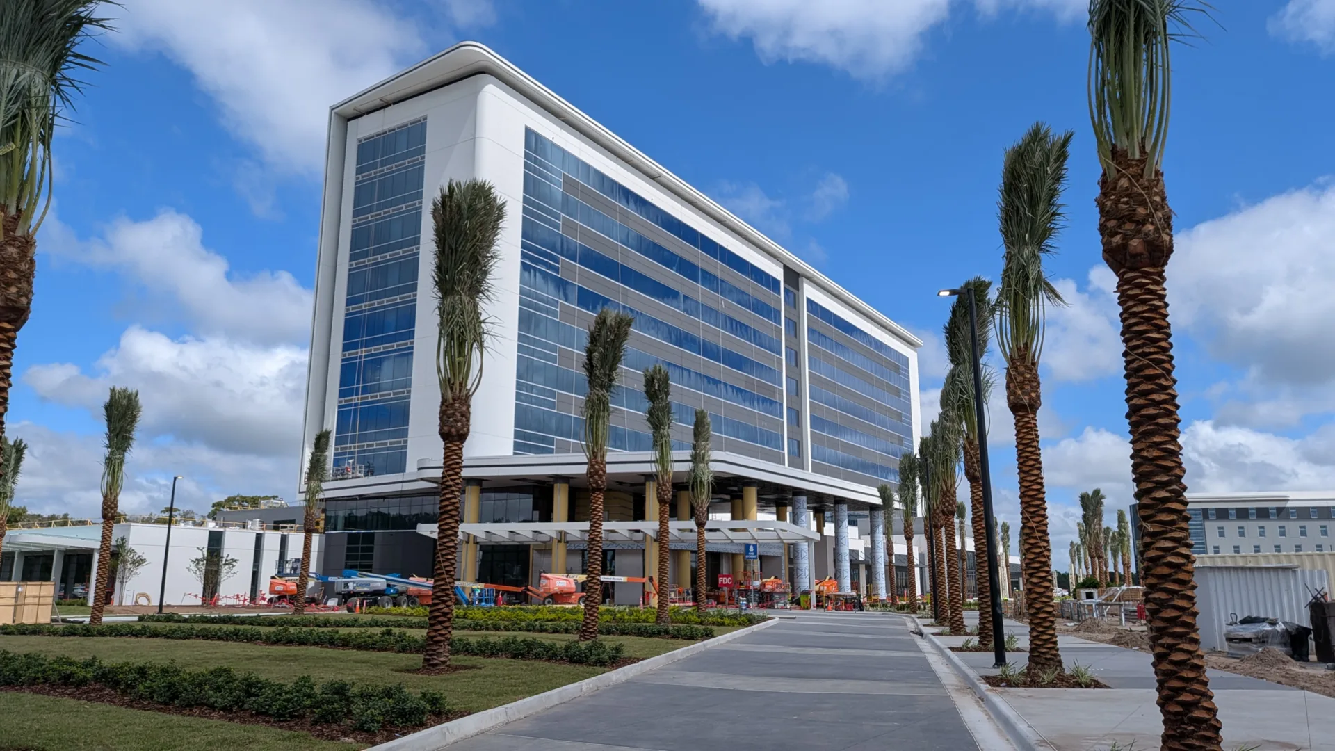 Ground-level, exterior view of a pathway leading to the entrance of hospital building, with palm trees along both sides of pathway and blue sky with white, puffy clouds in background
