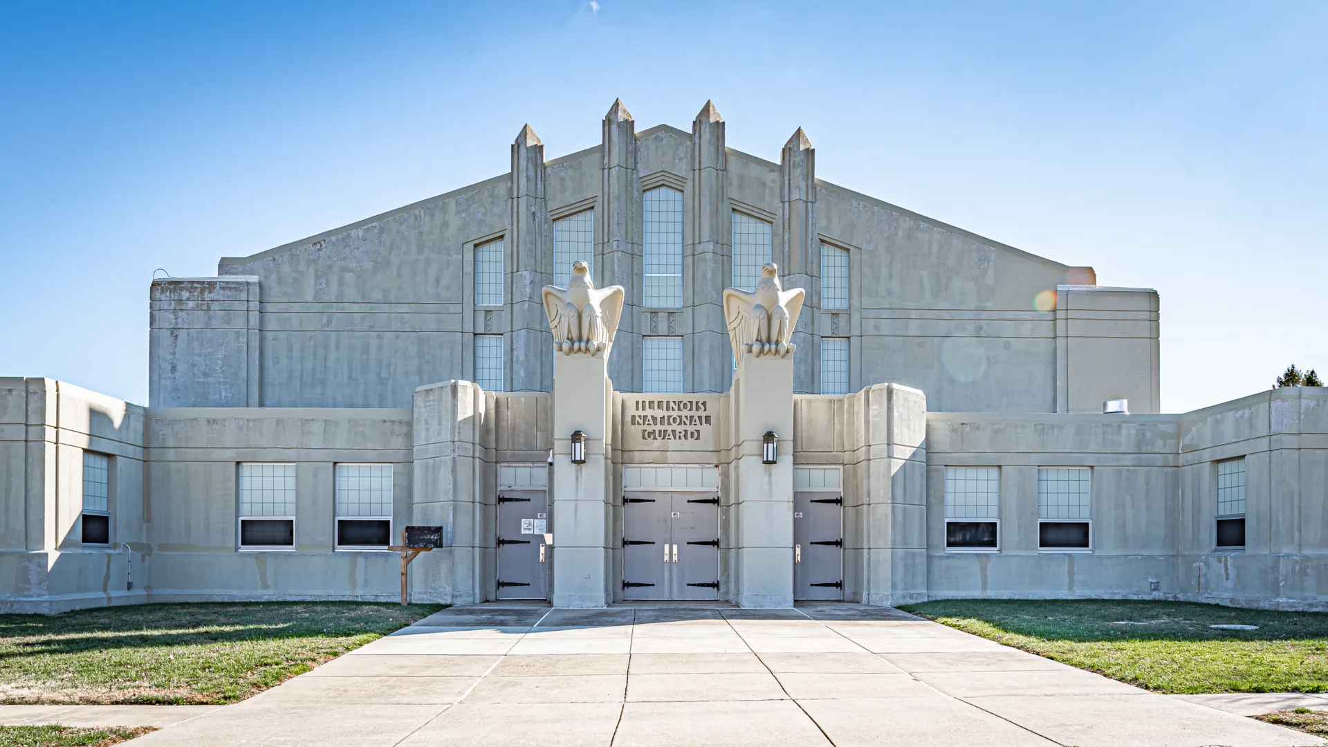 Exterior photo of a concrete building with walkway leading to front entrance flanked by columns with eagles on top. Illinois National Guard carved in concrete above main door
