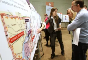 Woman with long, brown hair wearing dark suit pointing to large display board with map on it as several people on right look on