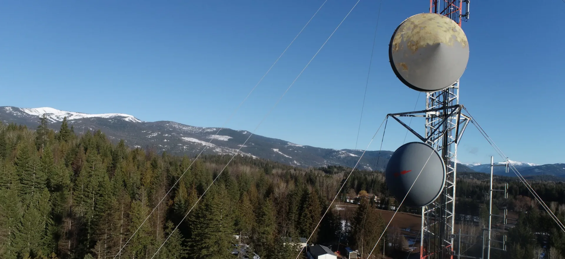 Drone photo of a telecommunications tower with mountains and pine trees and blue sky in background