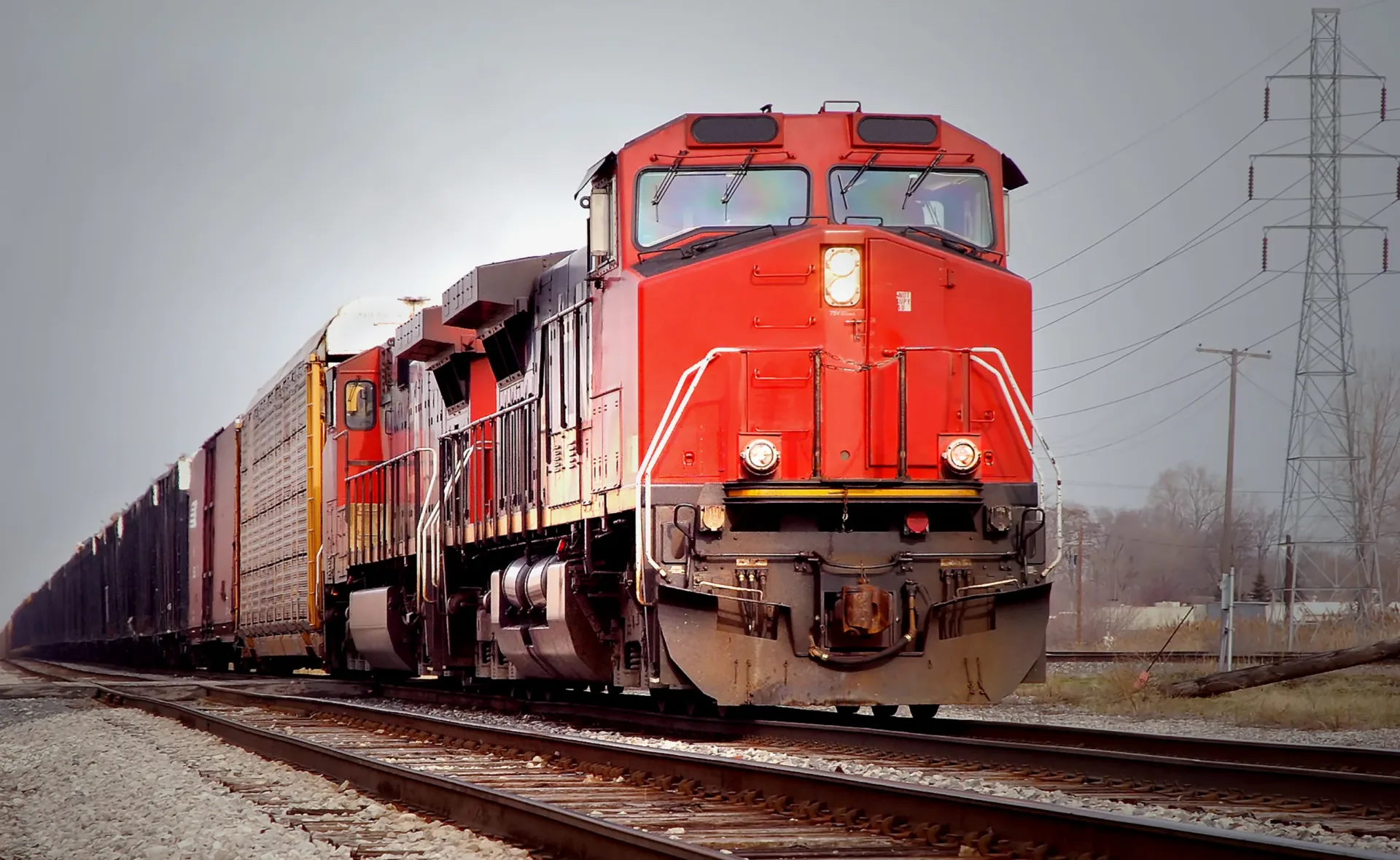 Ground-level image of a train engine pulling a train down one set of railroad tracks with a second set in the foreground along with gravel; gray sky and power lines overhead.