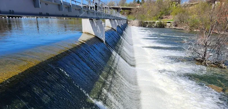 Side view of dam with water cascading down a ramp into a pond at the bottom; trees at right