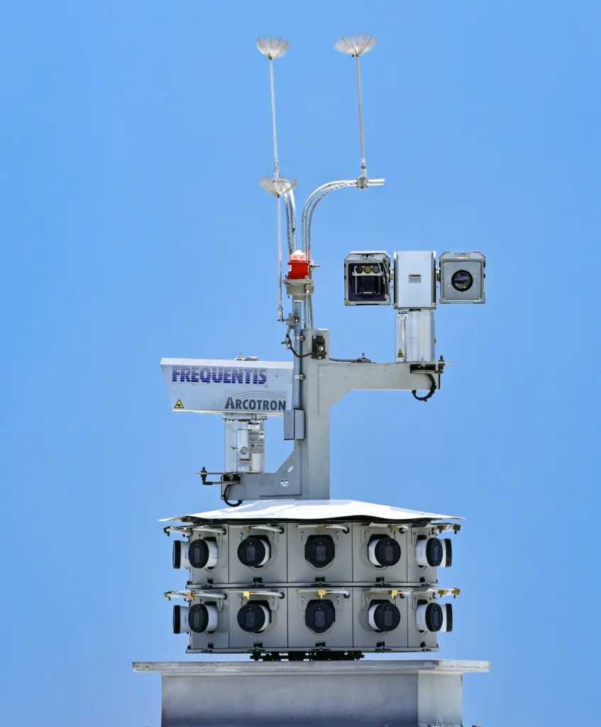 close-up photo of a double-camera array featuring two rows of cameras lined up around the base of a circular tower against a blue sky. Frequentis Arcotron printed on arm extending from camera