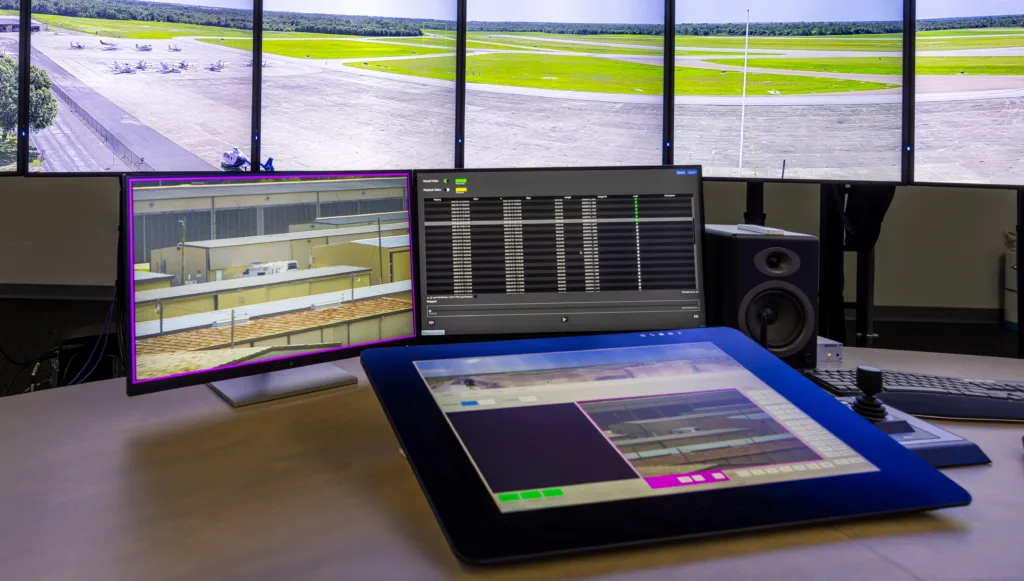 Close-up photo of two computer monitors and a large tablet on a desk with five large wall-mounted monitors in the background showing an airfield with planes at left