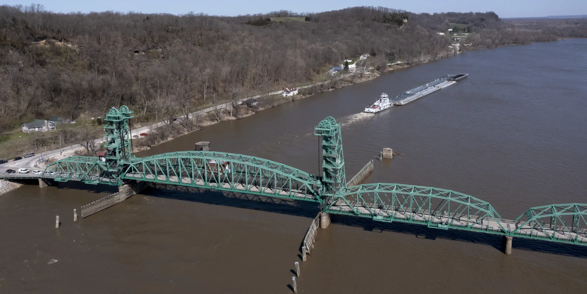 Aerial photo of teal-colored lift bridge on brown-toned river with barge in background and trees and roadway at left