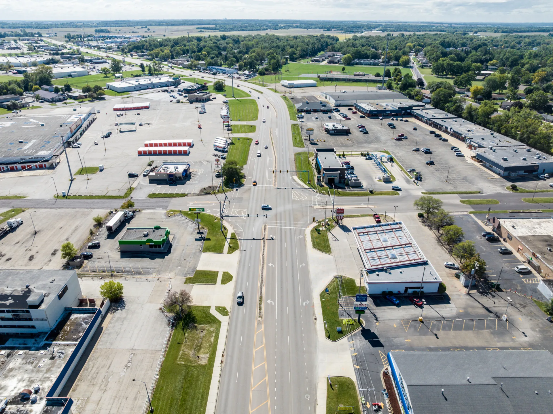 Aerial photo of a five-lane roadway curving to left, surrounded by buildings, parking lots and trees.