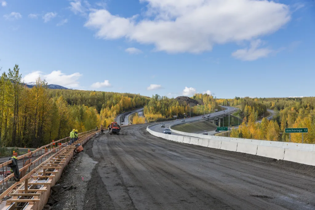 Roadway under construction with workers at right and cars passing at right on other side of concrete barrier; lots of yellow and green trees and some mountains in background