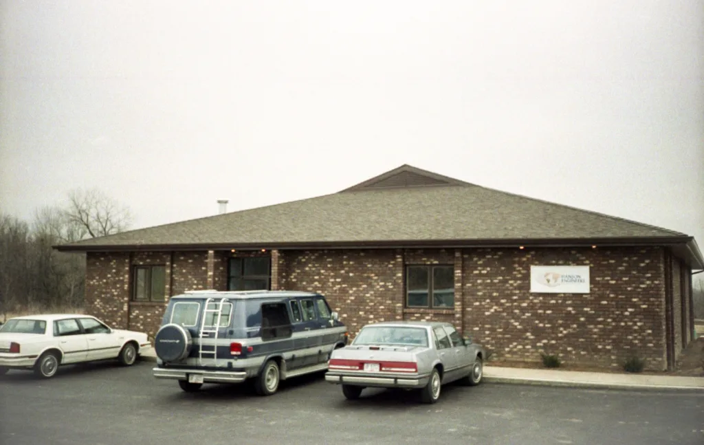 Exterior photo of single-story, brick building with Hanson Engineers sign at right and two cars and a van parked in front