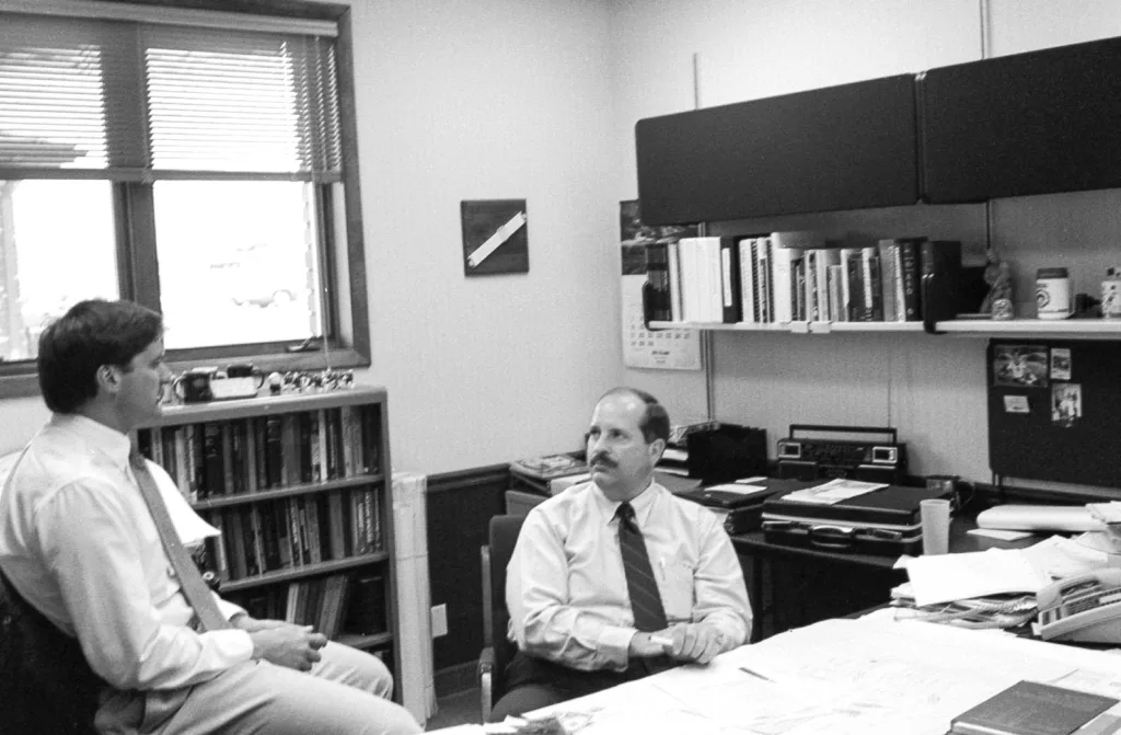 Two men talking in an office, both seated. Window in background at left and shelves with overhead bin at right; shelves lined with books; briefcase and boom box radio on desk under shelves.