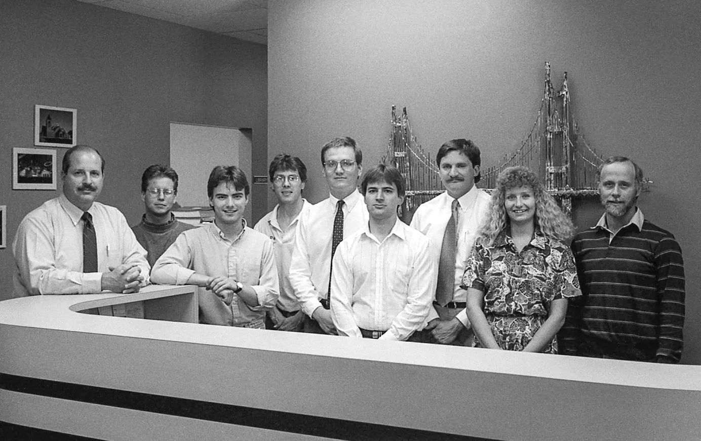 Black and white photo of nine people standing in a row behind a reception desk, most smiling, with a large piece of bridge-shaped artwork hanging on the wall behind them