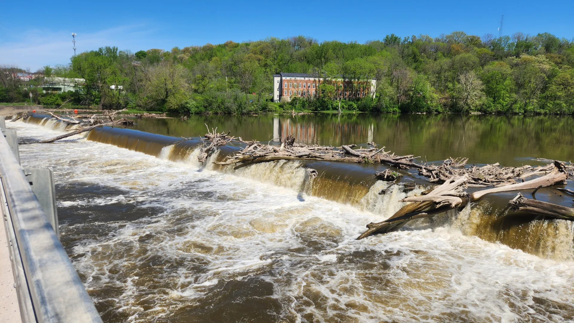 Water pouring over dam with tree branches being pushed over the edge; building in background surrounded by trees; water in center and foreground; choppy water at lower center and in foreground, with smooth water at right and in upper center