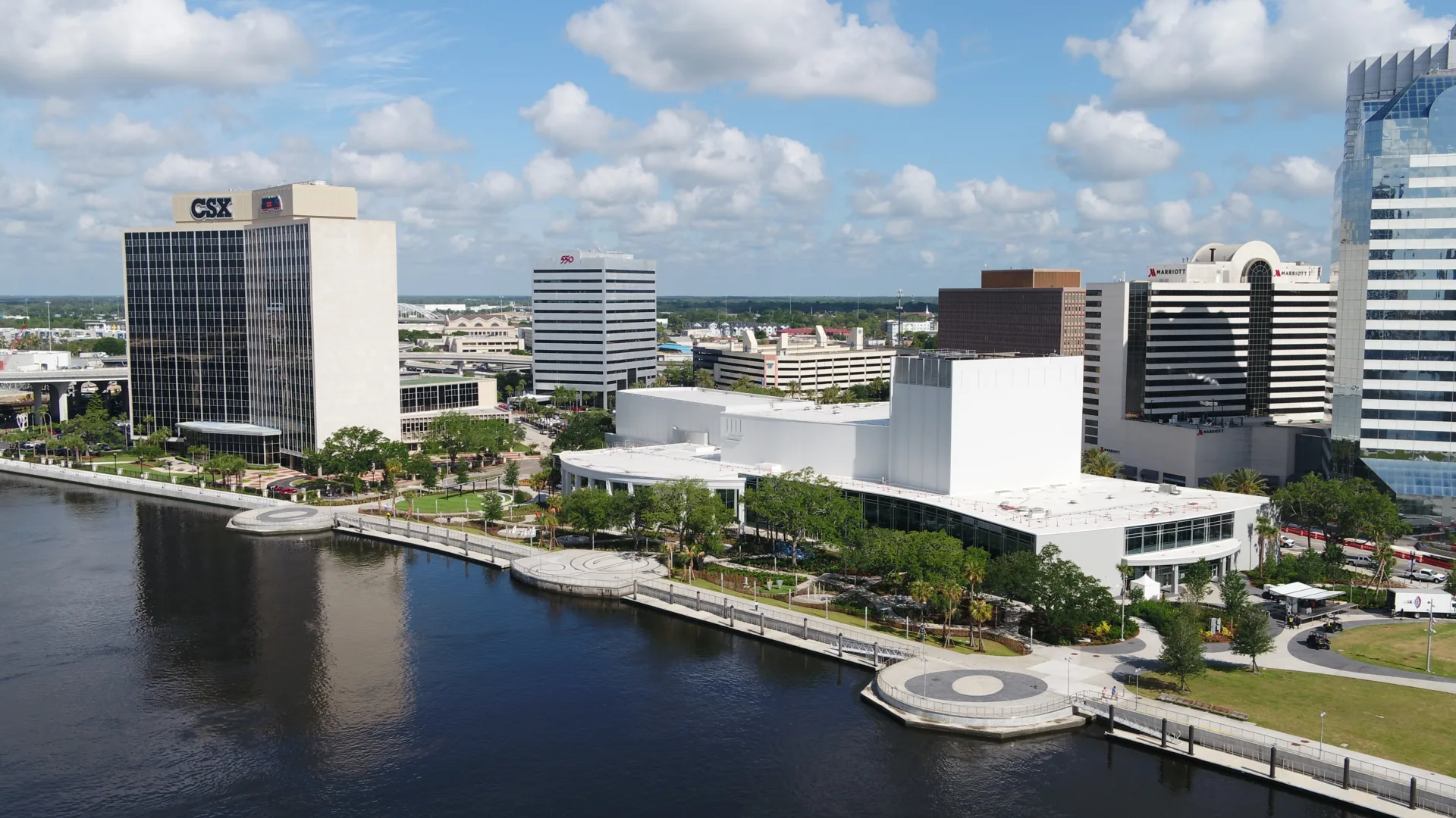 Aerial of a park along a riverside, with a musical note at center and city buildings in the background