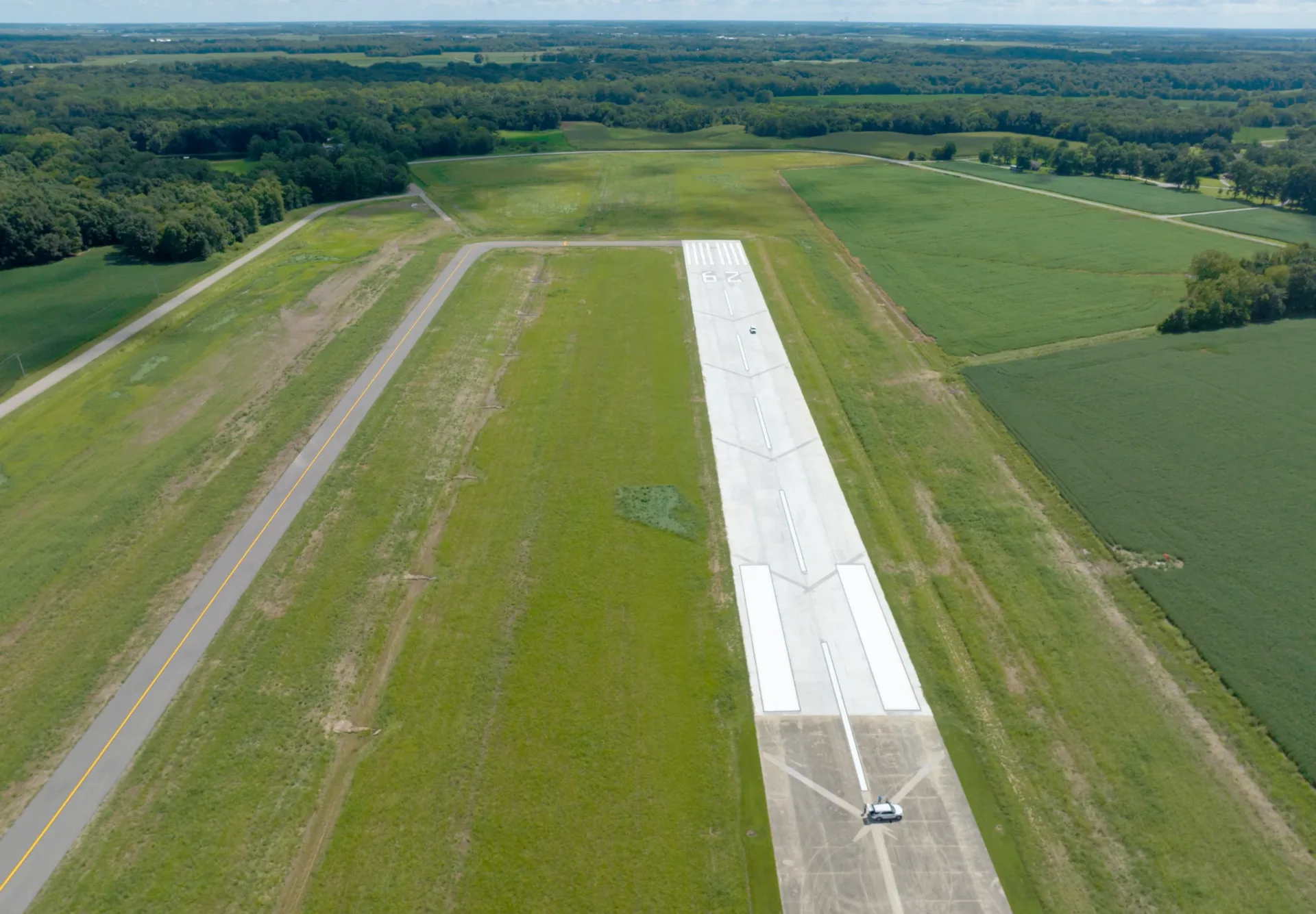 Aerial view of an extended runway with a taxiway at left turning to meet the end of the runway; grass and trees surrounding airfield