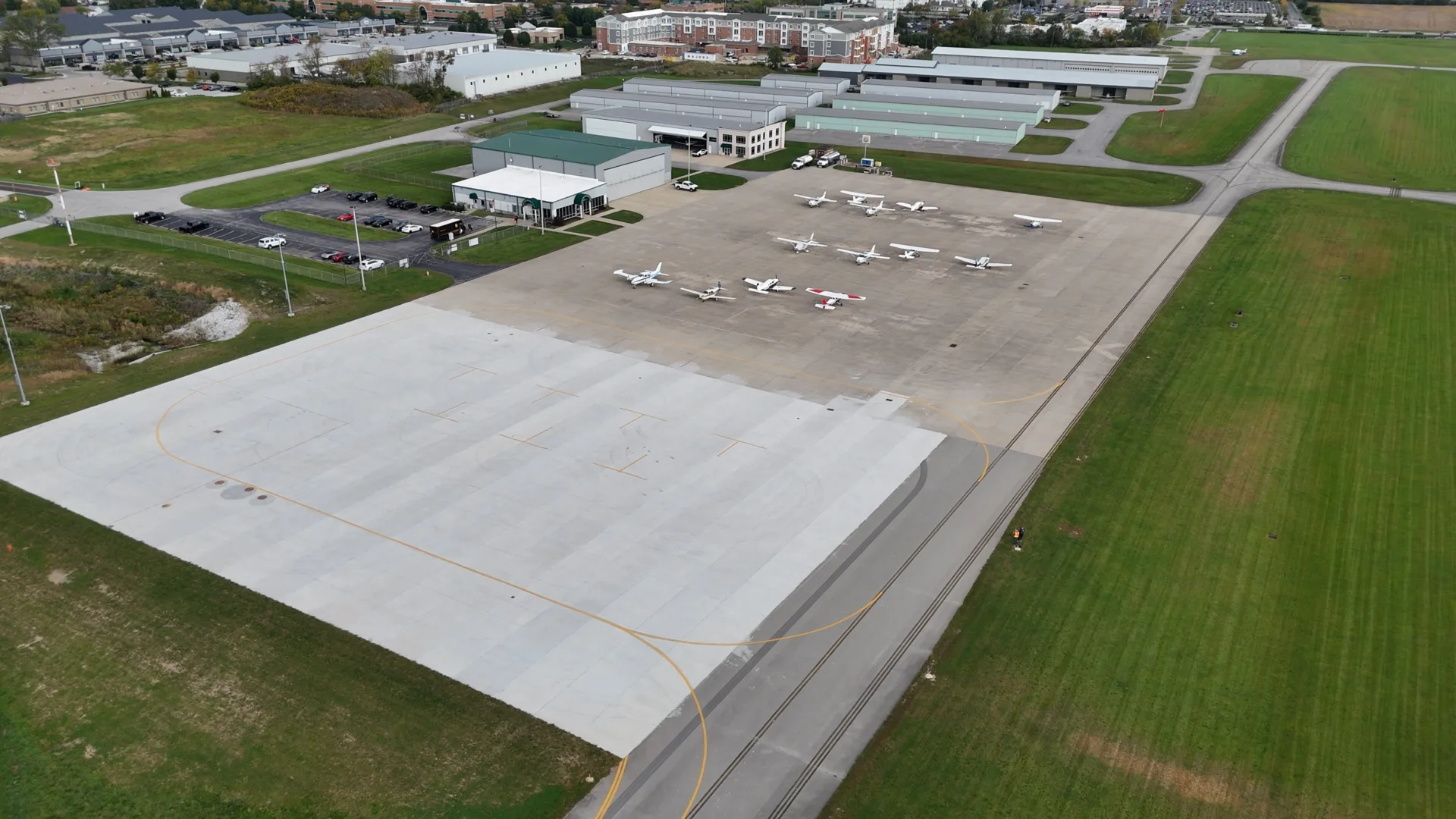 Aerial view of airport showing extended apron with half white and half darker pavement; 12 small aircraft parked on darker half; hangars and terminal building in background