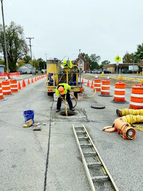 Construction on city street, with worker leaning over manhole holding hose extending down into manhole; orange construction barriers on either side and truck in background
