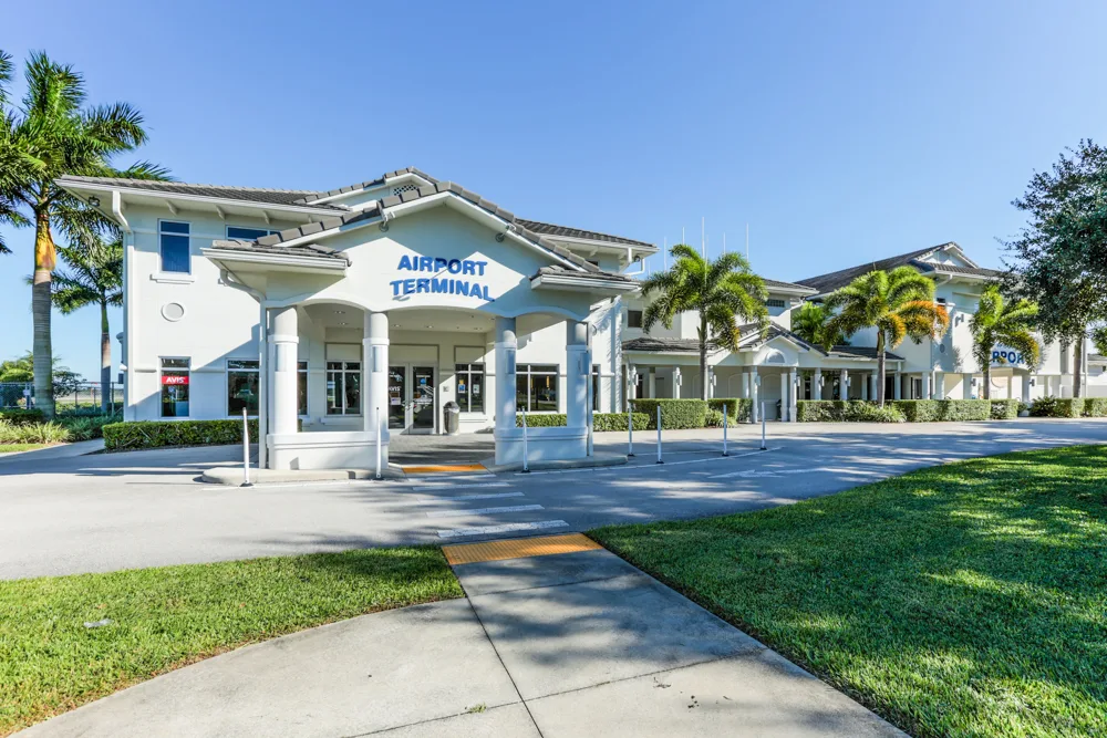 Building exterior with "airport terminal" on front of entrance canopy; other parts of building extending to the right with palm trees in front and sidewalk and grass in foreground