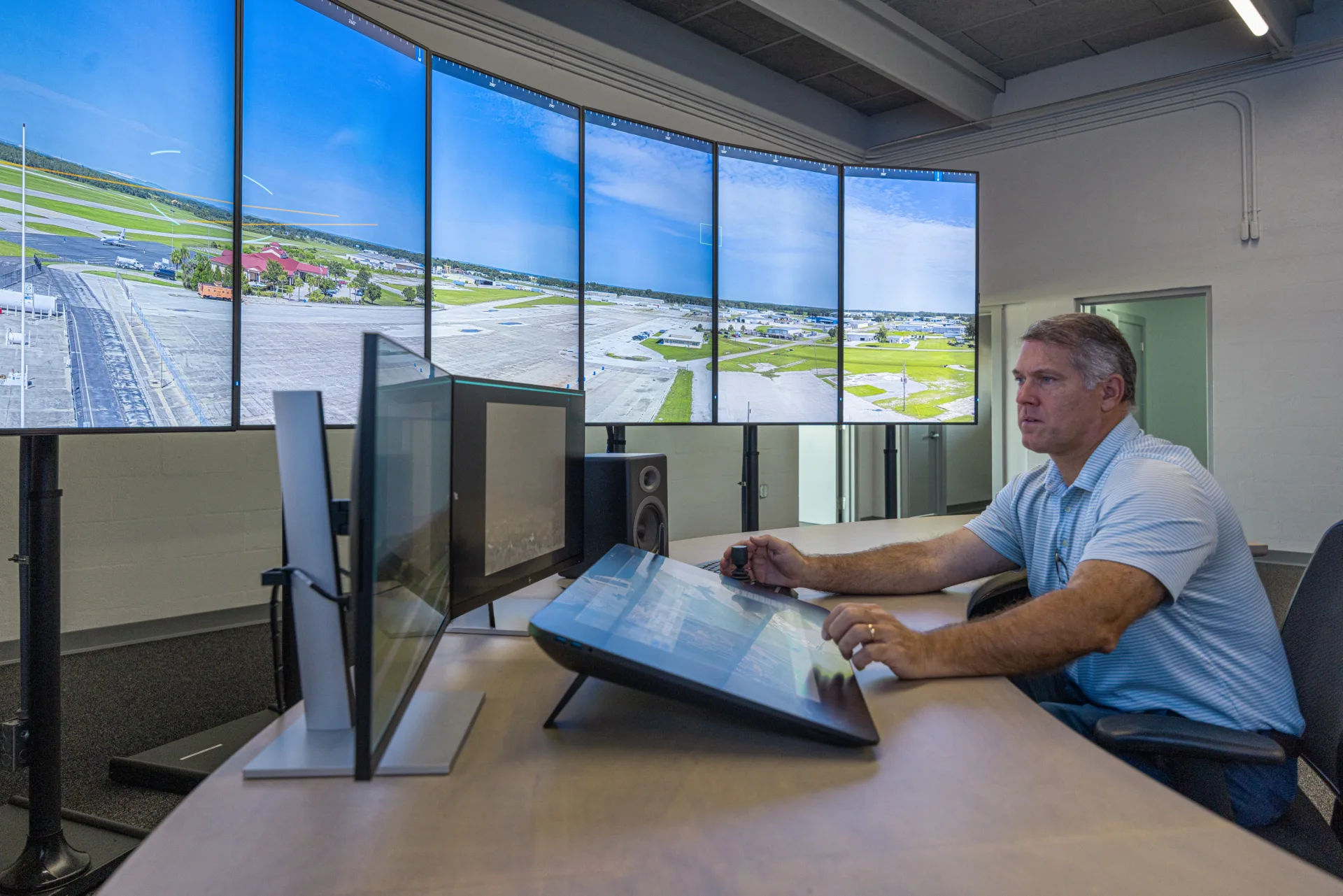 Man in striped shirt sitting at desk in front of computer equipment with row of large monitors showing airport runway at left and two sets of opened doors in background
